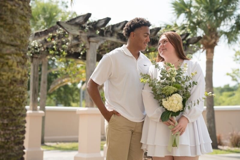 Happy interracial couple at St. Augustine Courthouse wedding ceremony standing outdoors under pergola, bride in white dress holding bouquet of white flowers and greenery, groom in white polo shirt and khaki pants, surrounded by lush tropical landscaping