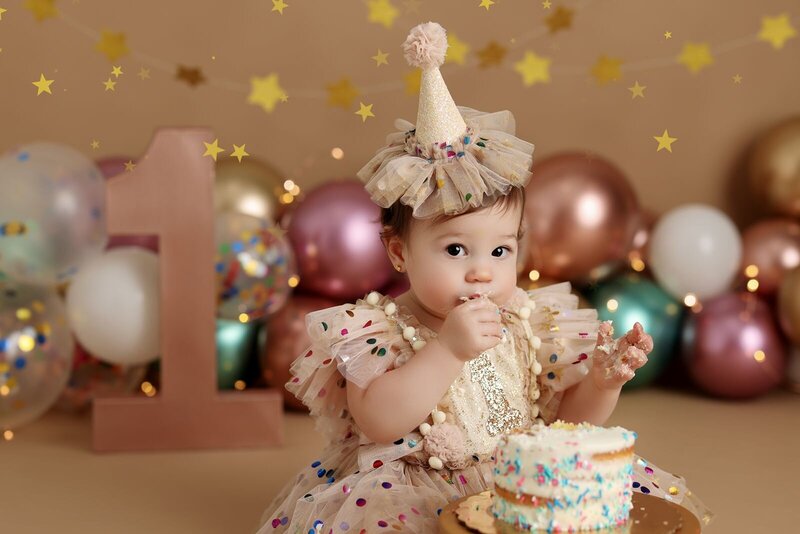 A baby girl celebrates her first birthday, dressed in a festive confetti-covered dress and matching party hat. She is joyfully tasting her cake, with a backdrop of colorful balloons and star decorations, creating a lively and cheerful scene.