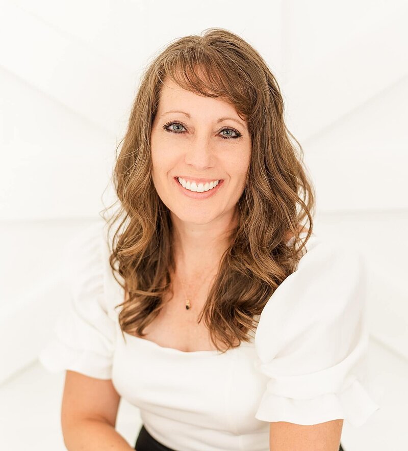 Jennifer Hall smiling and sitting by a white fireplace in a portrait studio.