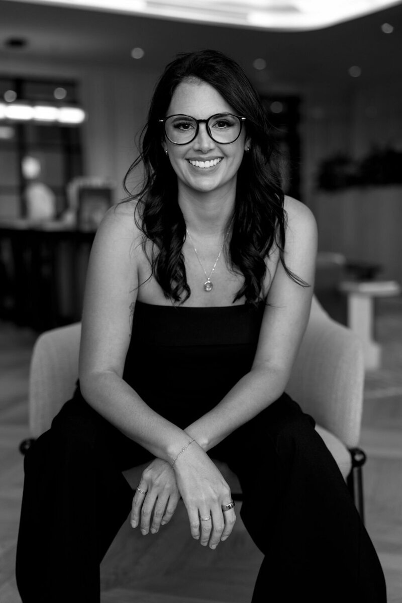 Raquel Bickford, founder of La Perle Retreats and ROQUE Events, smiles confidently in a black-and-white portrait, surrounded by elegant florals. Photo by Logan Alexander.
