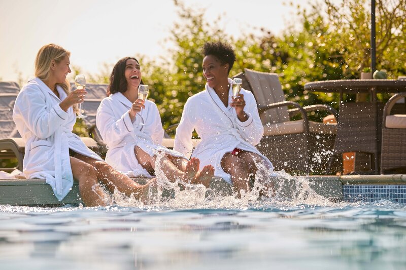 Three ladies laughing with their feet splashing in a pool, holding glasses with champagne