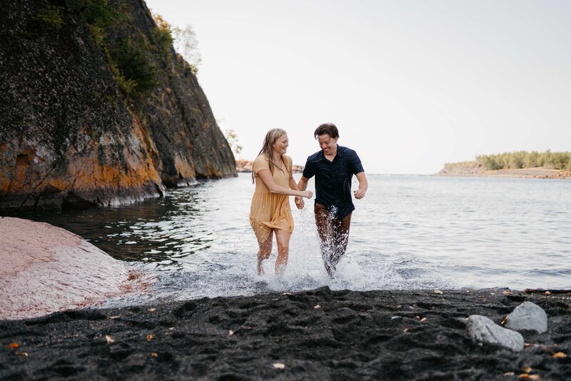 A couple running through lake superior at black sand beach - the perfect location for engagement photos on the north shore