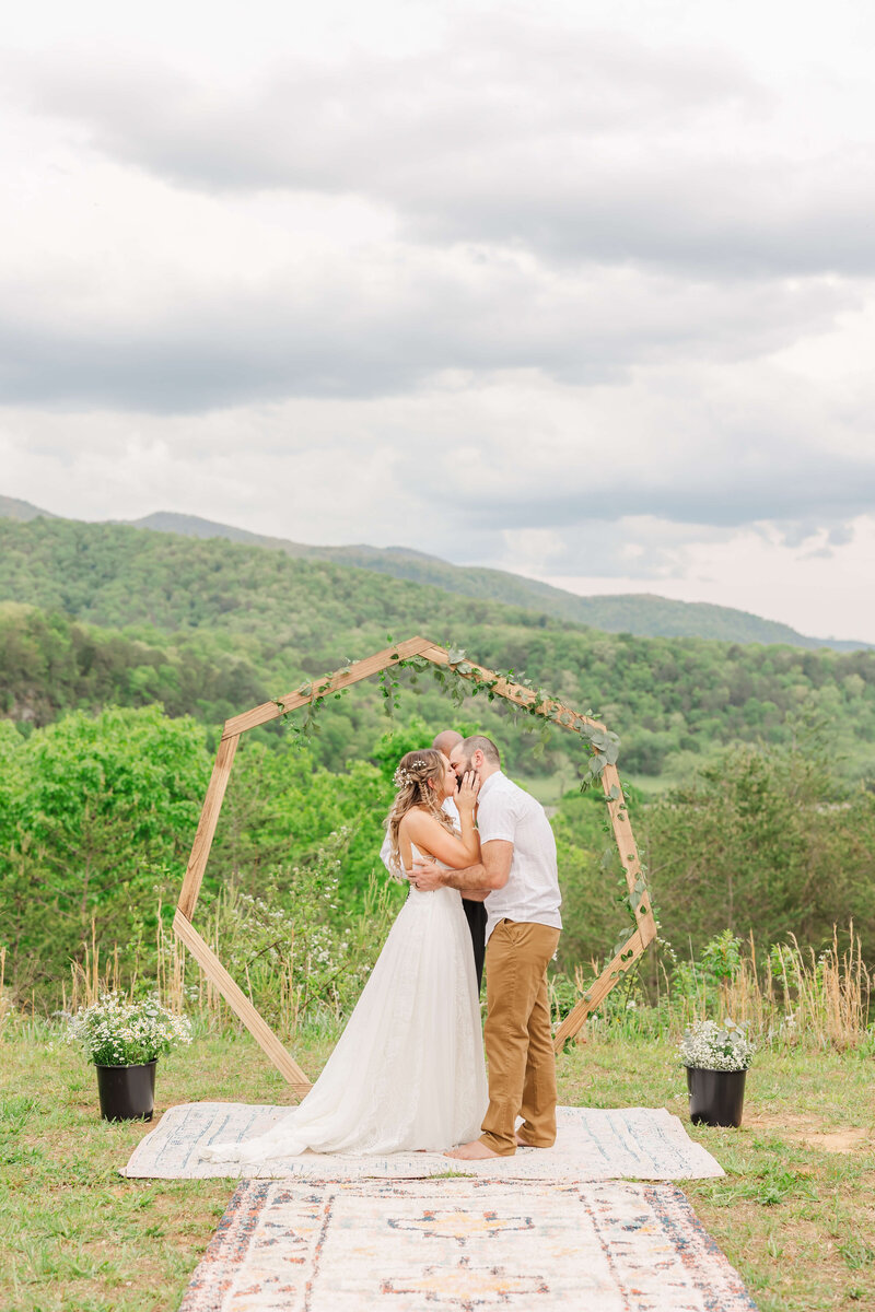 Bride and groom share a kiss during their outdoor mountain elopement ceremony under a wooden arch, captured by Destination Wedding Photographer.