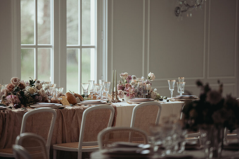 Light-filled reception setup with cream linens, florals, and white chairs surrounding a regular foldable rectangular table.