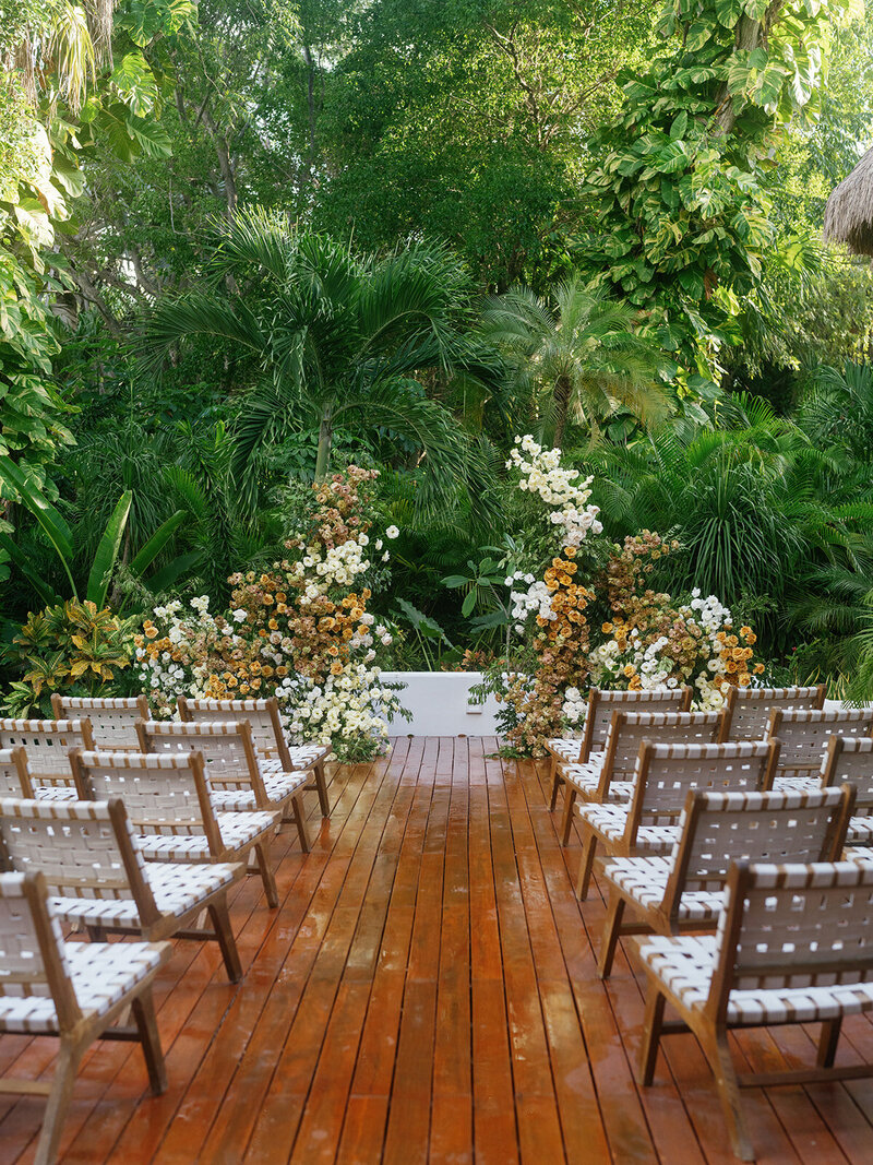 Diverse greenery provides a backdrop for a simple floral altar and wood plank wedding aisle between woven chairs
