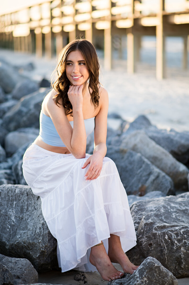 high school senior girl sitting on rocks near a pier on the beach in New Smyrna, FL