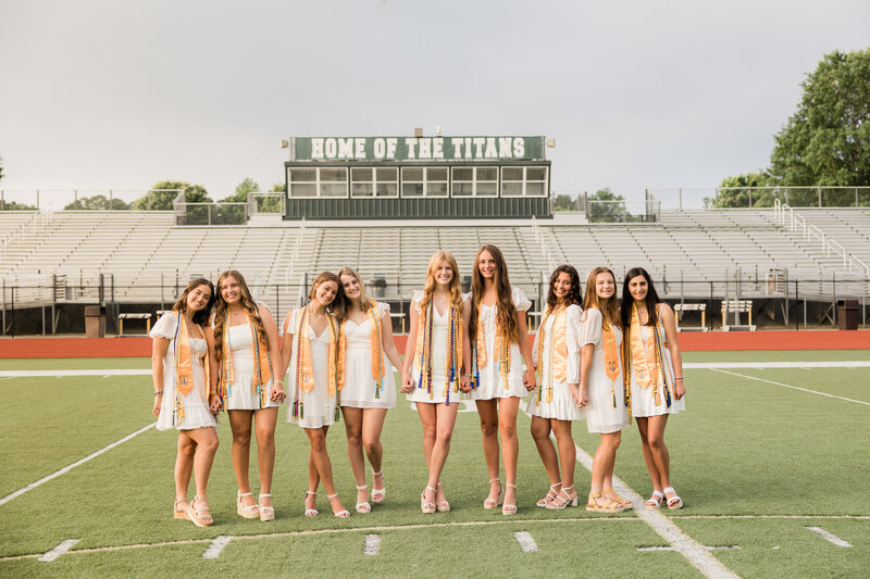 Group of females posed and smiling on a football field in graduation gowns.