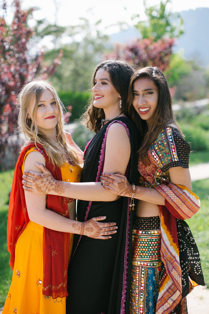 Three women dressed in vibrant Indian-inspired outfits embracing and smiling outdoors — colorful cultural portrait session with henna and traditional details