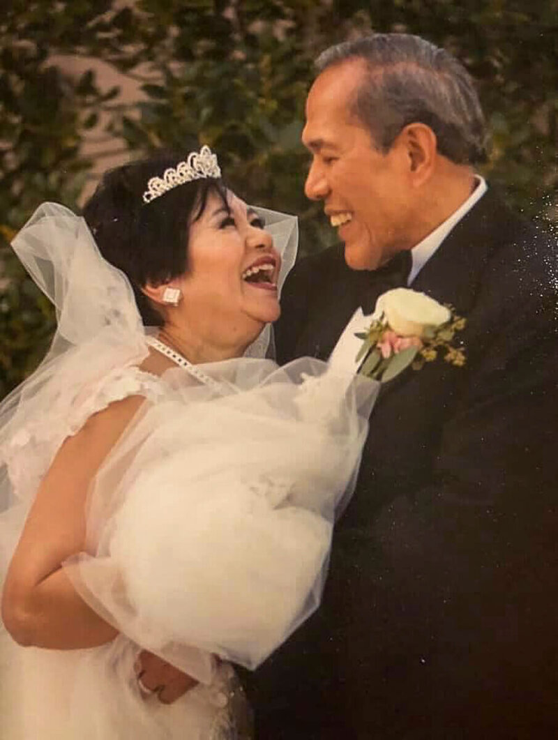 Nita and Pe Tin at their 50th wedding anniversary vow renewal, Nita in a beaded lace gown and tiara with full veil, Pe in black tuxedo, smiling and embracing one another, showing their enduring love and faith.
