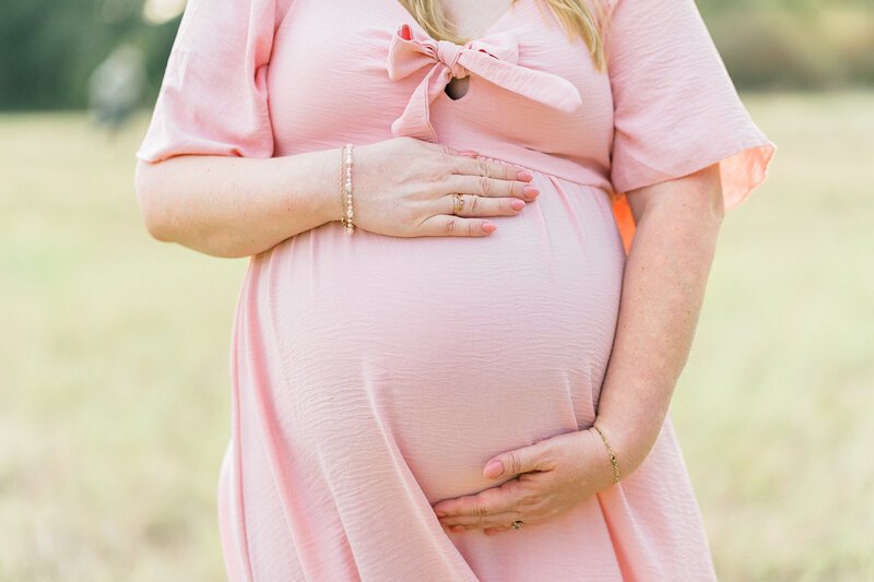 close up photo of a woman in a pink dress holding her pregnant belly during her photo session with a Cedar Park photographer.