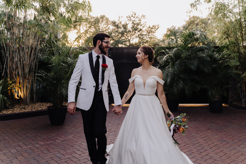 Bride and groom hold hands while looking at each other and walk towards the camera. Groom is wearing a white suit jacket and had a red rose boutonniere  and black tie. Bride is  wearing a white bridal gown and holding her bouquet of flowers. Photo taken by Orlando Wedding Photographer Four Loves