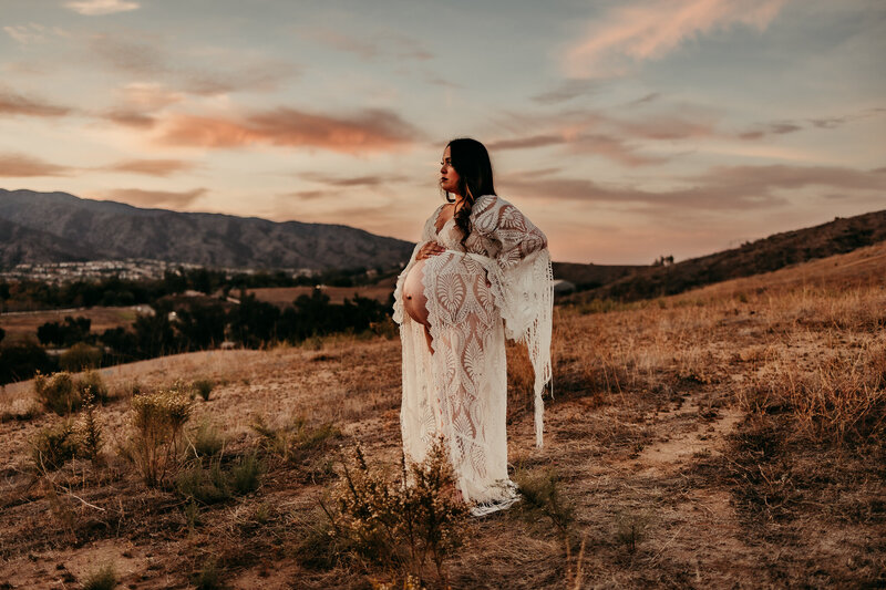 Pregnant woman wearing a white lace boho gown standing in a golden field at sunset with mountains in the background during a maternity photography session in Southern California

