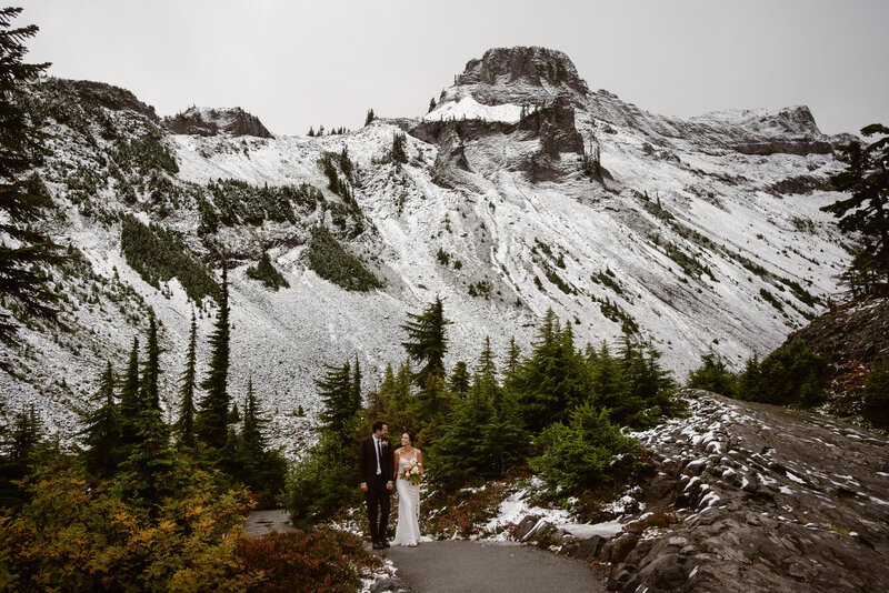 couple in wedding attire cheers with beer cans , sitting on ledge in front of mountain view