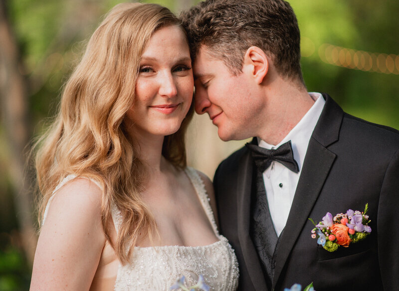 A bride with light blonde hair wearing a white wedding dress smiles gently while her groom places his forehead against her cheek