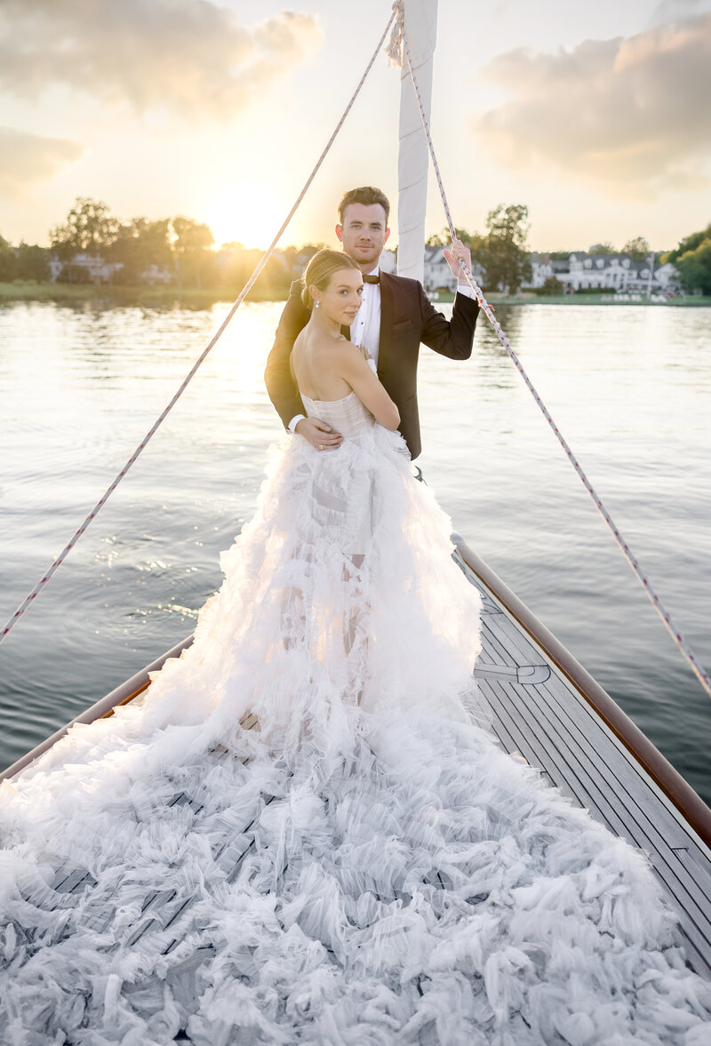A bride and groom stand on a boat at sunset. The bride wears a flowing white gown with a long train, and the groom is in a black suit. They embrace while looking at the camera, surrounded by calm water and a serene sky.
