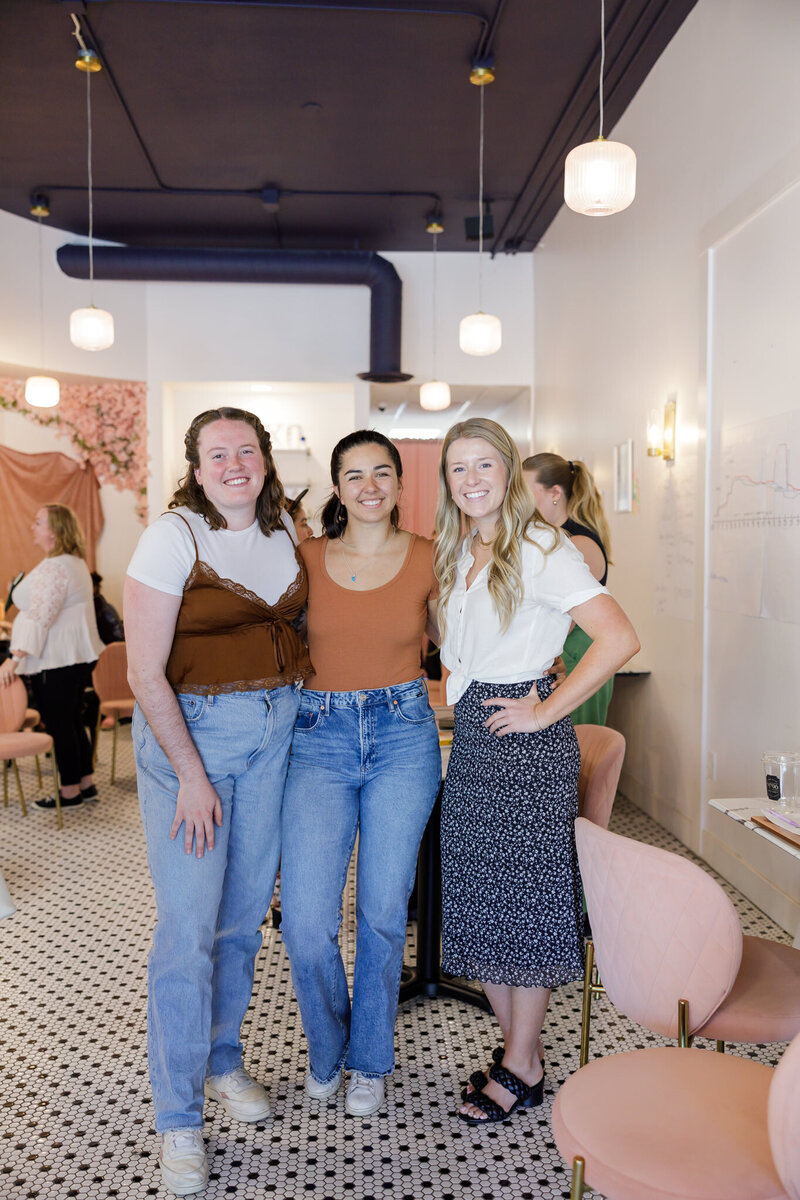 Three women stand closely together, smiling at the camera in a brightly lit, modern cafe in Seattle. The woman on the left wears a white shirt with a brown top, the middle woman wears an orange shirt with jeans, and the woman on the right sports a white blouse and patterned skirt. It's clear they are enjoying some networking time.