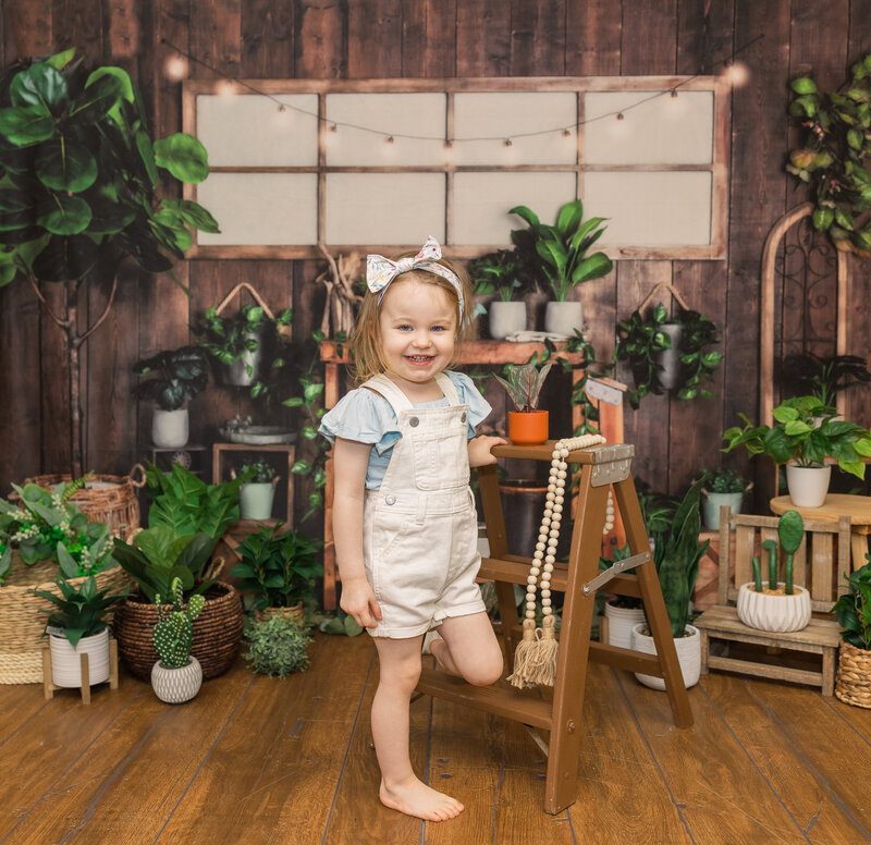 Frozen Moments by Kathy Photography | Small child in white overalls in a brown studio surrounded by plants
