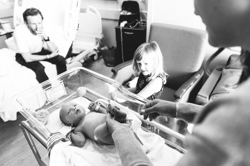 Hello & Co. Photography, family in a hospital room with big sister looking at a newborn baby in a bassinet