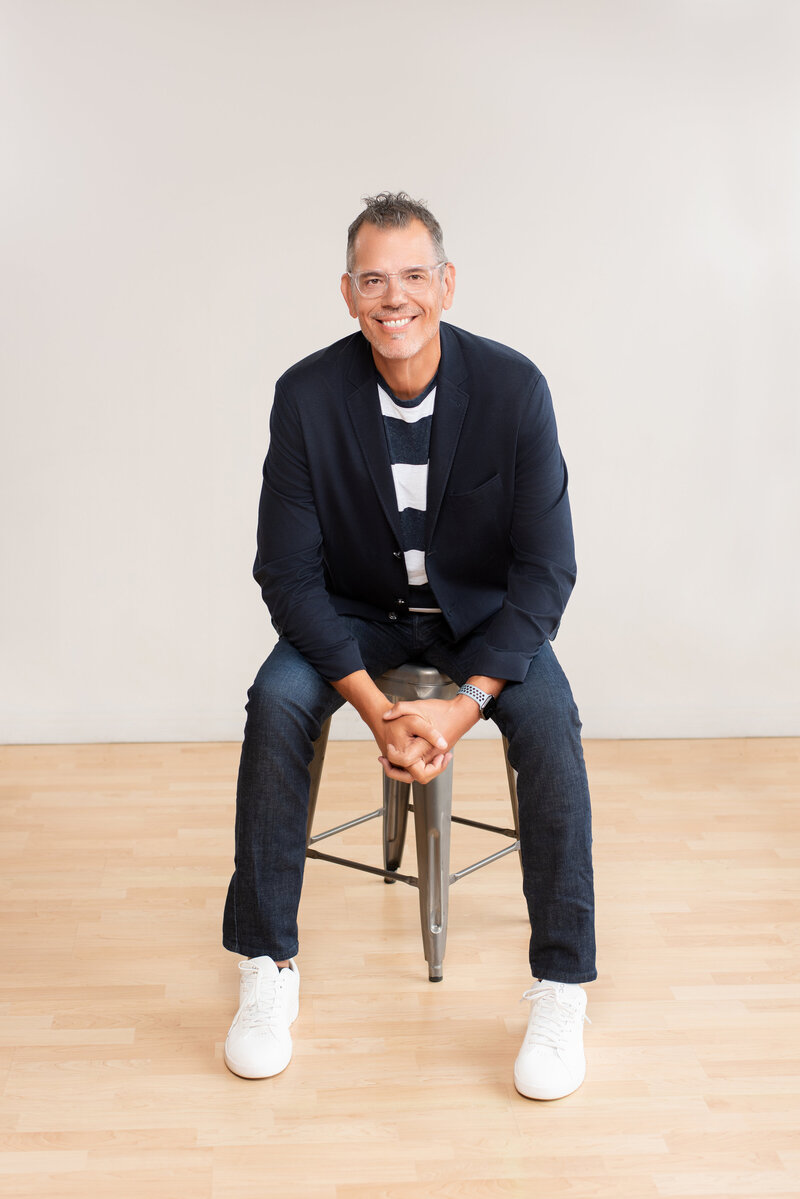 A tall man sits on a stool, dressed casually to take a professional headshot.