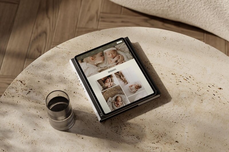 Tablet lying on a textured stone coffee table beside a glass of dark coffee, showing a newborn photography website gallery with close-up portraits of sleeping babies in soft neutral tones.