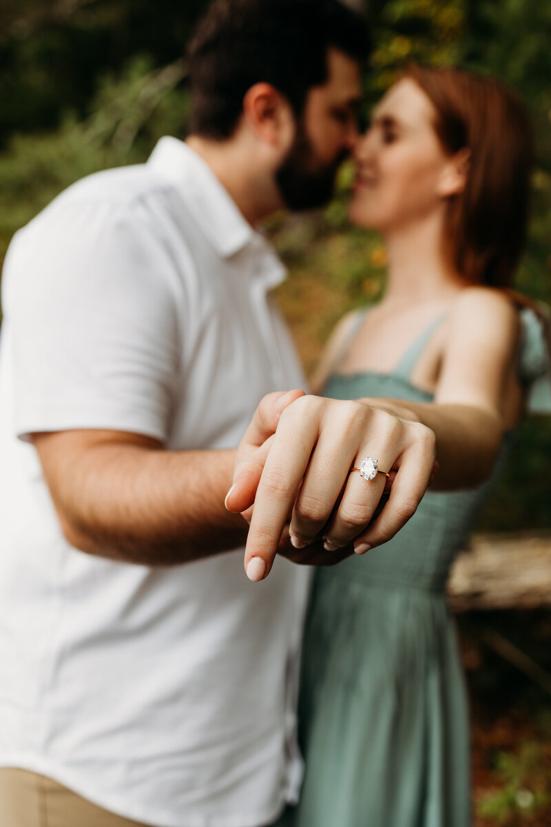 An engaged couple leans in for a kiss while holding out the woman's ring hand to show off the ring