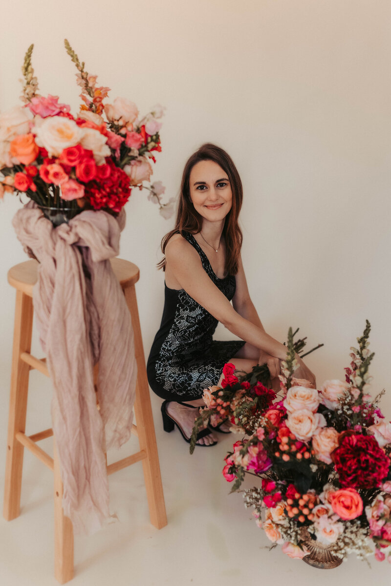 Woman in black dress sitting holding pink and white flowers in hands surrounded by floral arrangements against a cream colored backdrop.