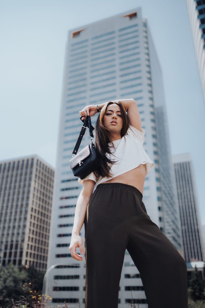 Woman in white crop top and black pants holds an arm over her head holding a small, black purse in front of large skyscrapers.