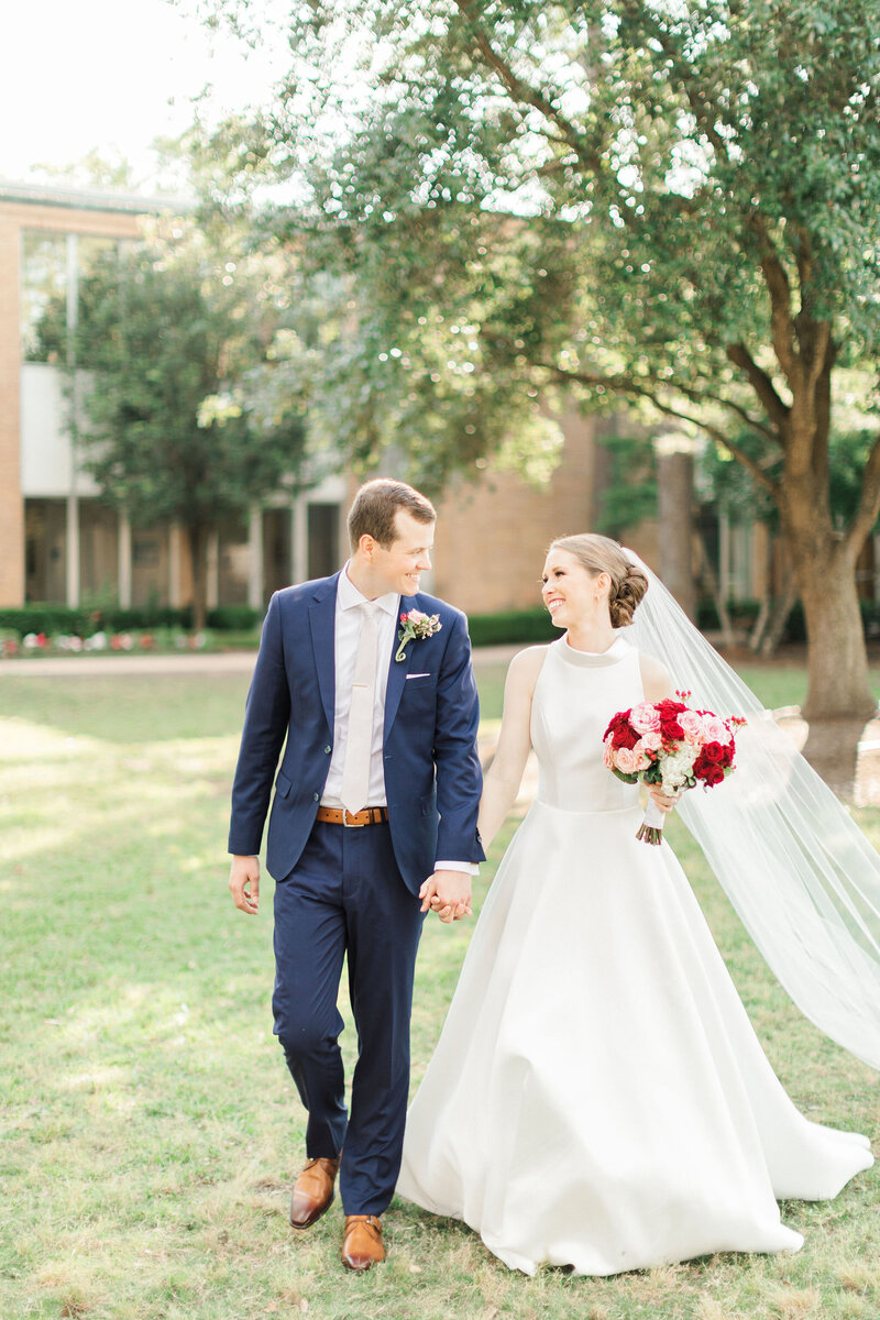 Steve is wearing a navy blue suit and looks at his wife with love and holds her hand as they walk across the lawn at Duchesne Academy before their reception