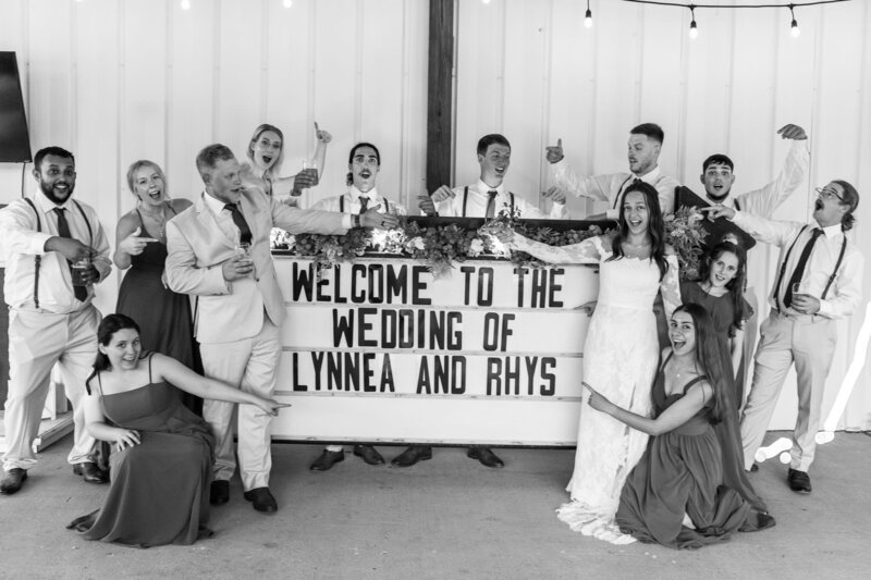 A group of excited guests crowds around a marquee sign that reads "Welcome to the Wedding" during a laid-back Central PA celebration. The couple stands front and center, laughing with friends and family dressed in suspenders, suits, and dresses. The energy is lighthearted and rooted in community, with joyful chaos in the best way.