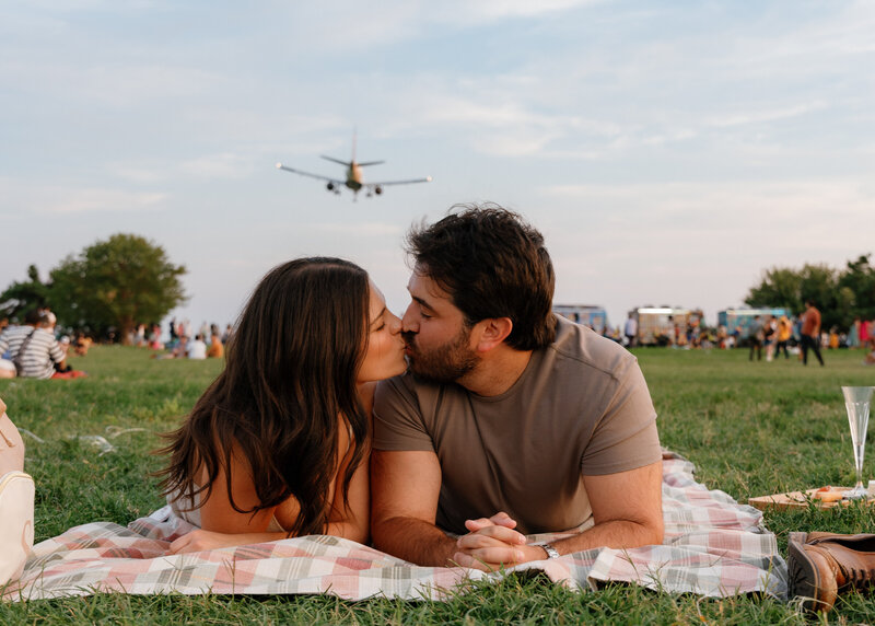 A couple lays on a picnic blanket as a plane flies overhead