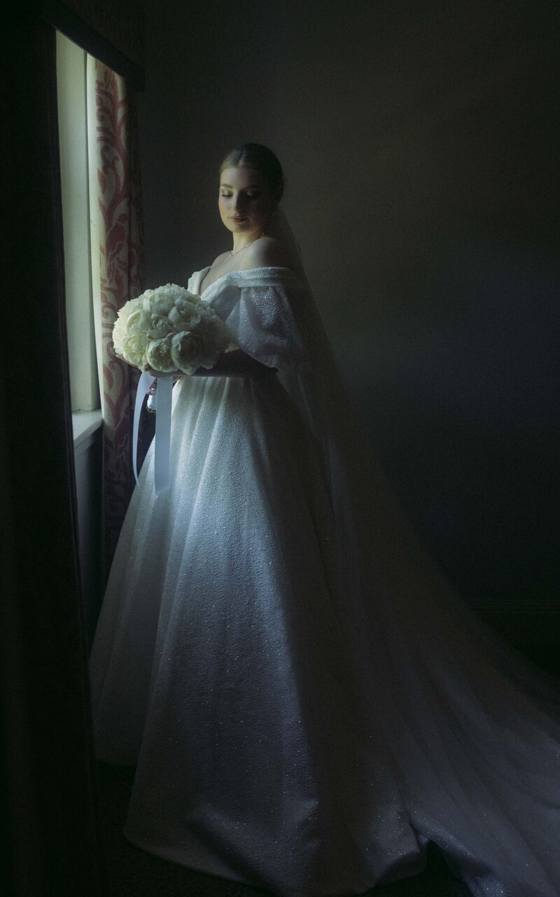 Bride standing next to the window with soft, natural light highlighting her beauty, creating a serene and elegant moment before the big day