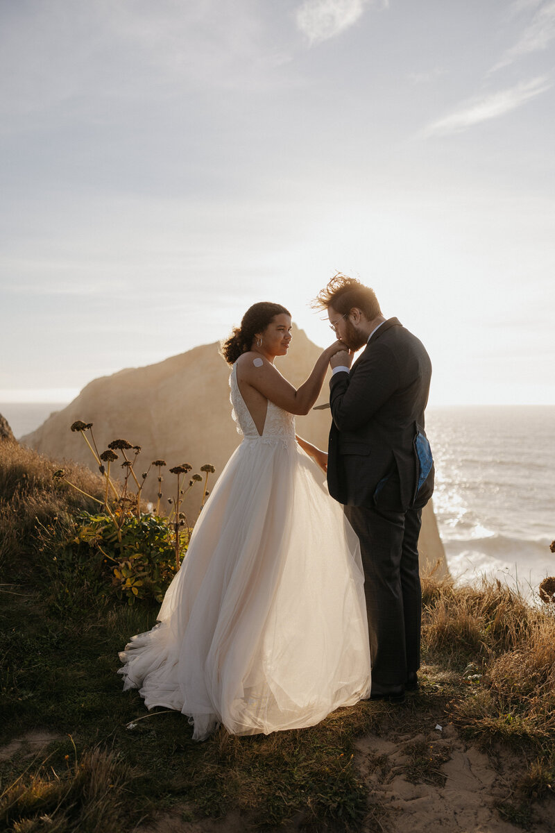 A newlywed kissing their partner's hand on an ocean cliff 