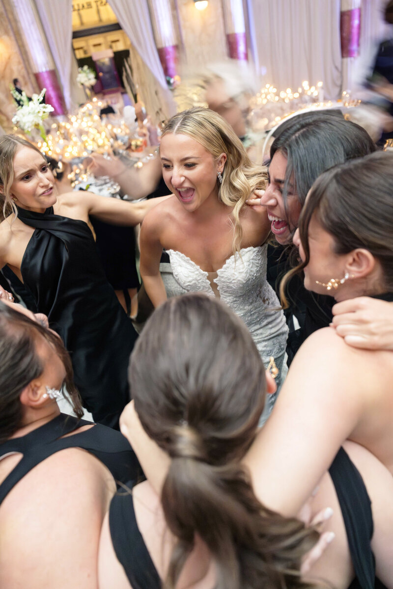 A bride in a white gown and bridesmaids in black dresses joyfully gather in a circle, arms around each other, smiling and celebrating indoors, with blurred festive lights in the background.