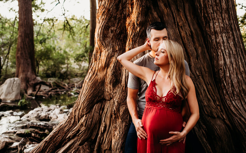 Maternity Photographer, husband and wife embtace, her hands on her belly before large tree in forest