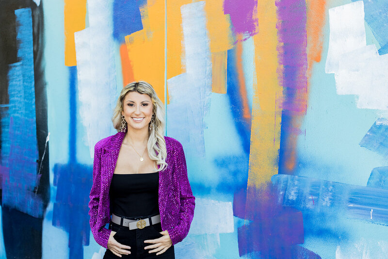 Smiling woman in a vibrant purple sequin jacket poses confidently against a colorful abstract mural in Columbia, SC. Her black outfit, gold belt, and statement earrings complement the bold, artistic backdrop. Small business branding photography in Columbia, SC, capturing modern, stylish personal branding for entrepreneurs and creatives.