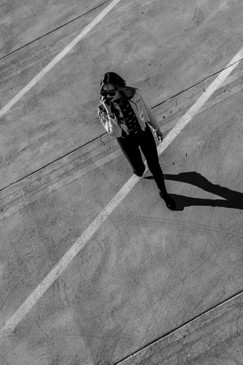 Black and white photo of a blonde woman, Heather Jiron, walking across a parking lot holding a phone 