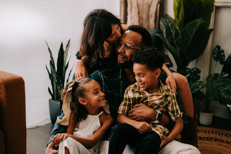 Family Photography, Family huddled around dad, mom is kissing husband while children laugh.