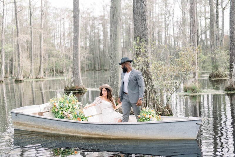 Bride and groom in a rowboat with flowers holding hands bride wearing a white lace dress and groom in a  gray suit and hat