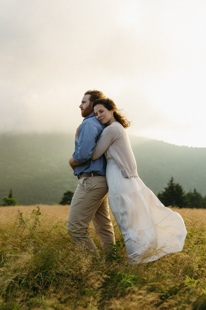 A woman hugging a man while the sun sets behind them and the Blue Ridge Mountains, captured by Andrea-Marie Photography