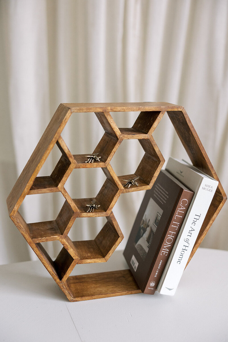 A wooden hexagonal bookshelf sits on a white surface, holding two books titled "Call it Home" and "The Art of Home." Small bee figurines are placed within some of the hexagonal compartments. A light-colored curtain is in the background.