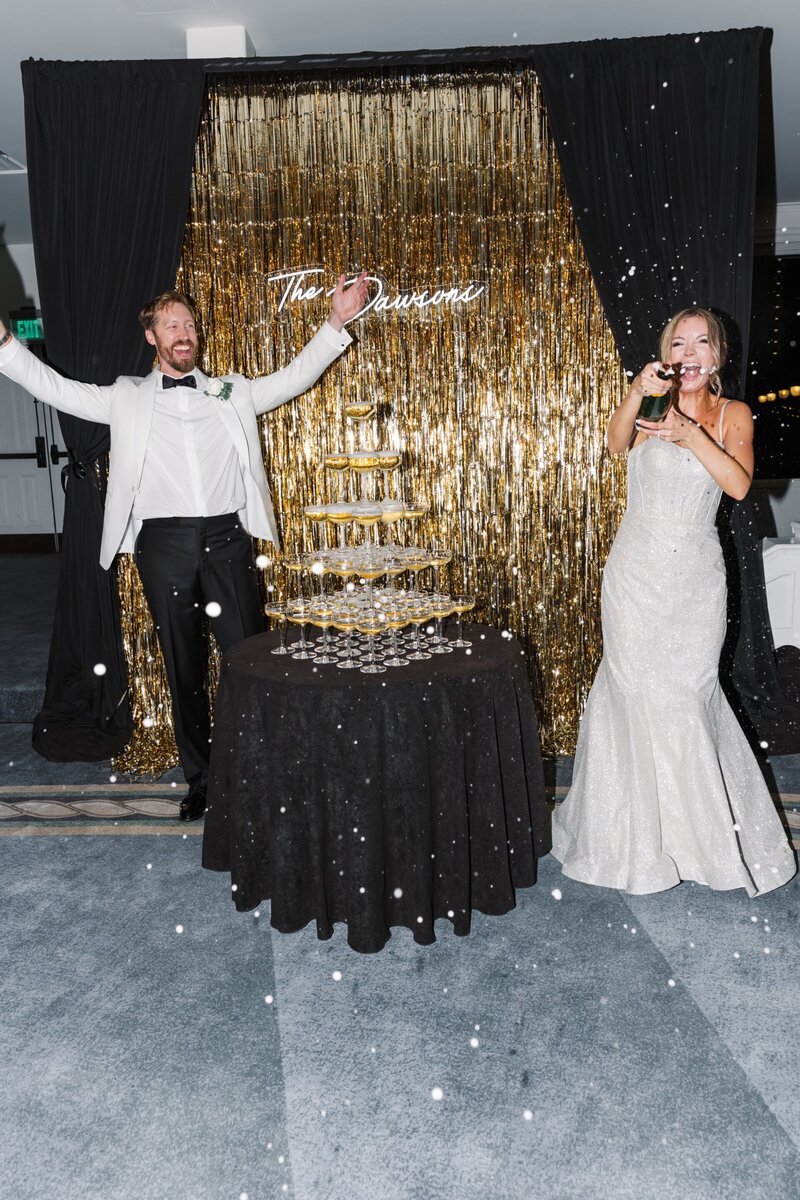 Two newlyweds celebrating by a champagne tower in front of a gold backdrop with a neon sign during their wedding reception at Hotel del Coronado. This wedding was planned and designed by Thater Made, a luxury wedding and event producer based in San Diego, CA.