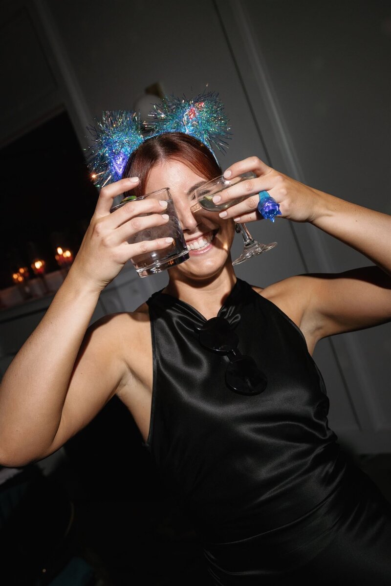 Woman in silk black halter dress wearing a bluish tinsel cat ear LED headband holding two cocktails over her eyes smiling at the camera