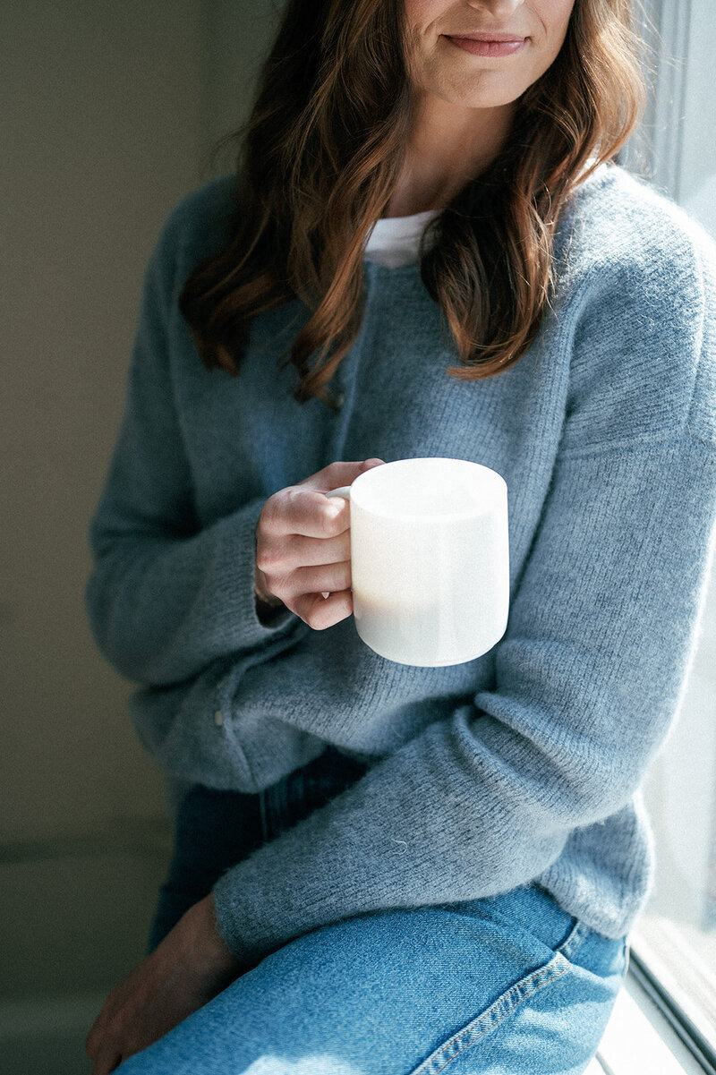 Woman in a blue sweater holding a white mug while sitting on a windowsill
