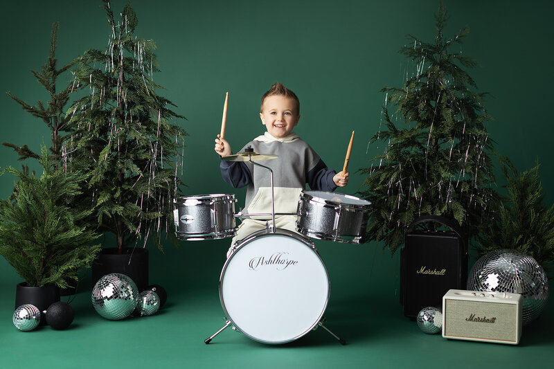 A smiling young boy plays a silver drum set surrounded by Christmas trees, disco balls, and Marshall speakers in a rockstar-themed holiday setup. Captured during a Dallas Baby Milestone photography session.