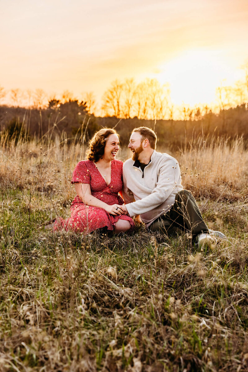 couple laughing together while sitting in a field at sunset for engagement session