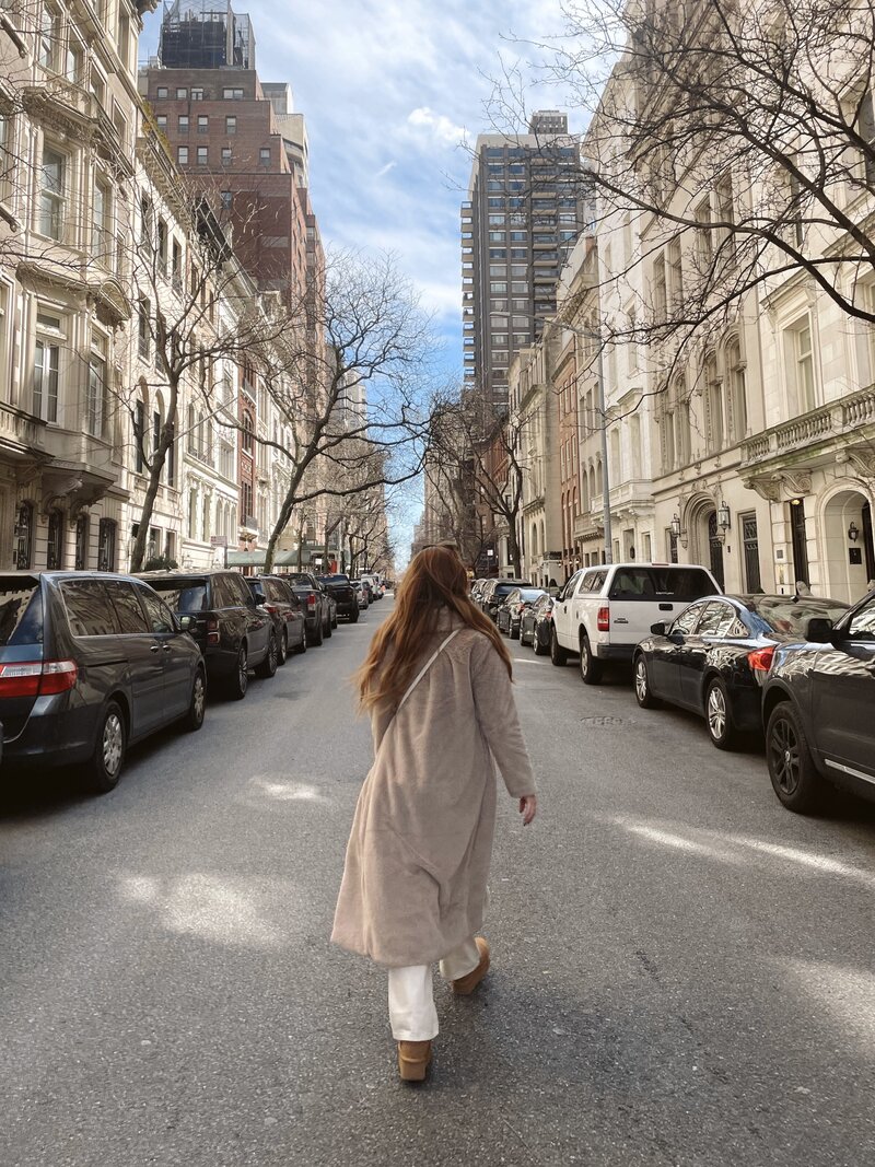 Woman walking down a quiet tree-lined street in Manhattan, New York City, surrounded by classic brownstone buildings and parked cars on a sunny day — representing mindfulness, city living, and calm urban balance.