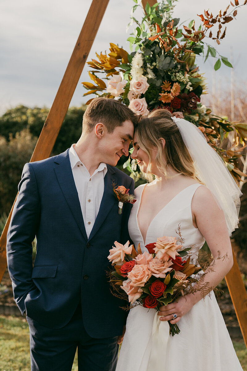 Close up of the bride and groom as they stand in front of their ceremony arch. Their foreheads are touching and they look blissfully happy