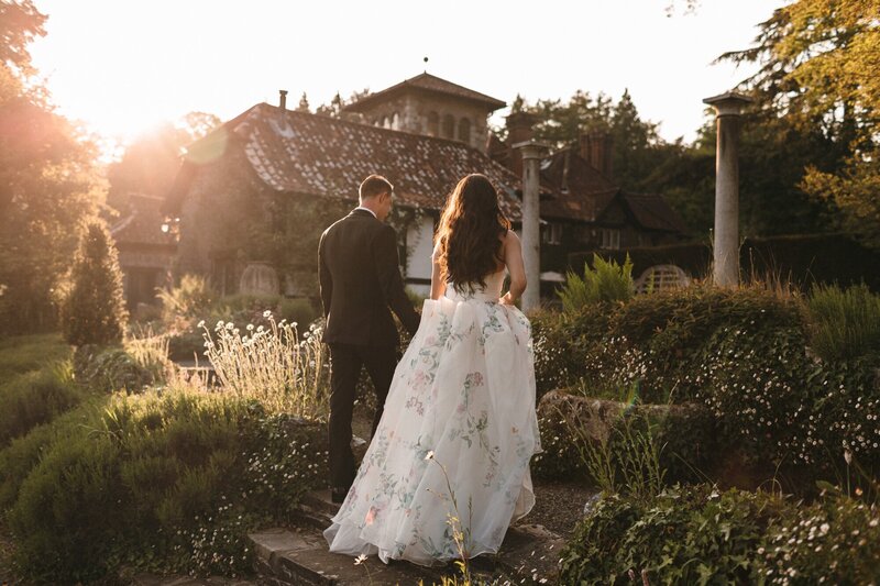 A bride wearing a floral wedding dress and her groom walk through daisies into the Italian Terrace area at their Devon wedding 