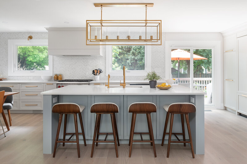 Large kitchen with large painted blue island with low-back wooden chairs, there is a rectagular cage light above. The kitchen has white counter tops, and herringbone while tiles along the wall