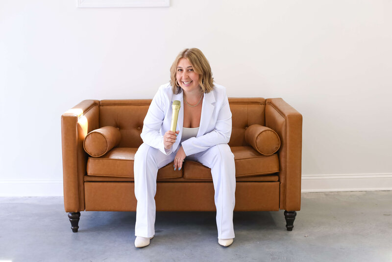 A woman in a white suit sits on a brown leather couch, smiling and holding a microphone, against a plain white wall.